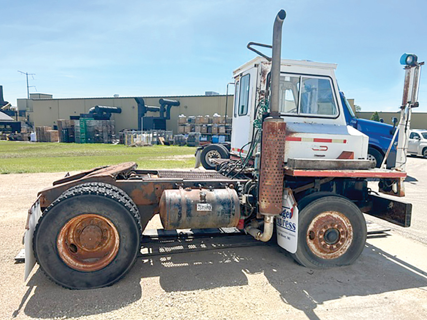 Truck chassis side view showing diesel fuel tank, exhaust stack, rear fenders, and white cab with blue accents; visible circular rims and heavy-duty tires.
