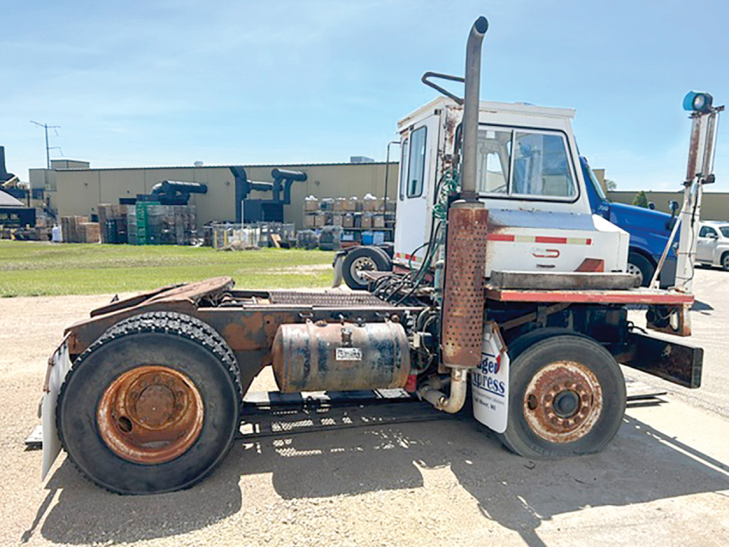 Truck chassis side view showing diesel fuel tank, exhaust stack, rear fenders, and white cab with blue accents; visible circular rims and heavy-duty tires.