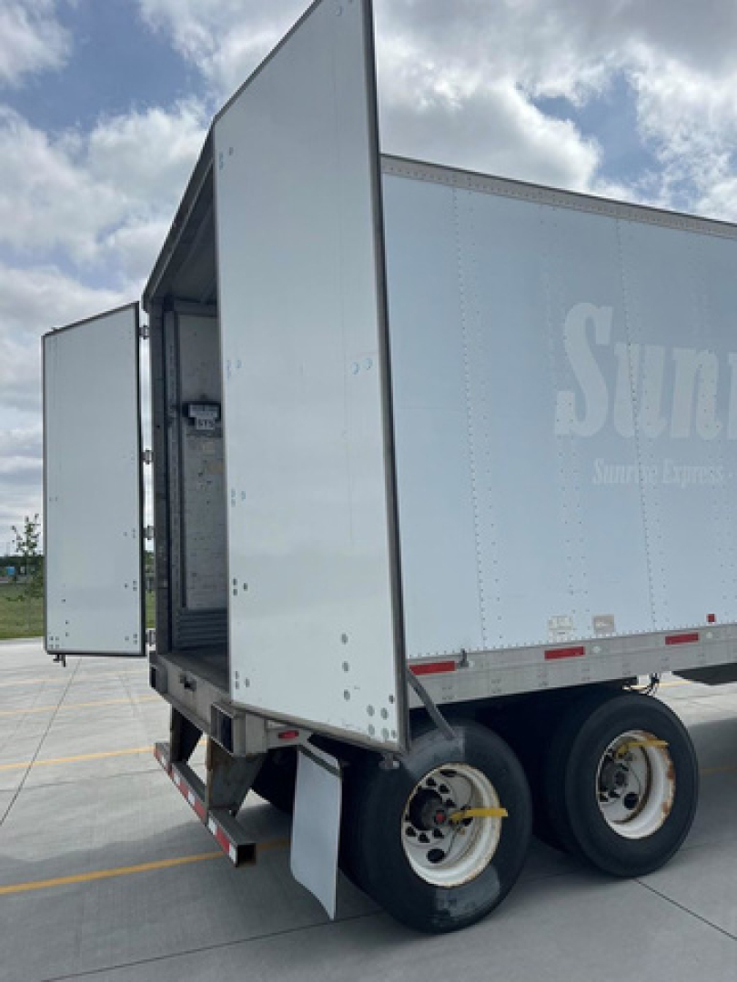 Truck trailer side showing open rear door and dual rear wheels, light gray box trailer with SUn logo fade, open loading area and interior showing metal frame.