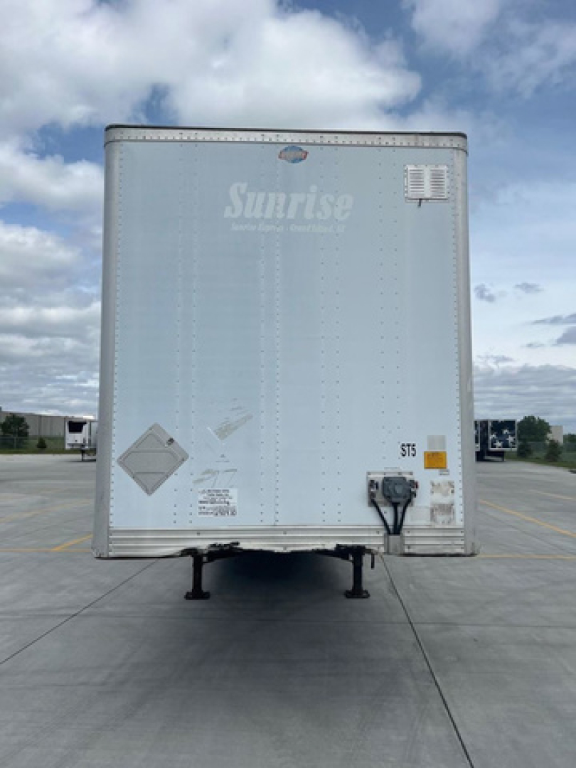 Front view of a light gray Sunrise utility trailer with a nose wheel, standing on a paved lot. There are warning placards and a vent on the front door.