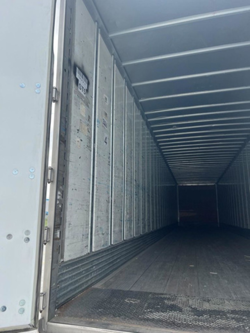 Interior of a trailer with metal sidewalls and a darkened long cargo area, entry from open rear door, visible riveted panels and floor.