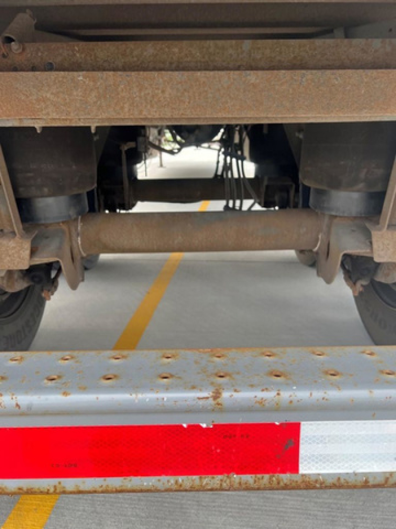 Underbelly of a utility van showing rusty chassis rails and cross members; rear bumper with white and red reflective strip; view toward the wheel hubs and drive line components.