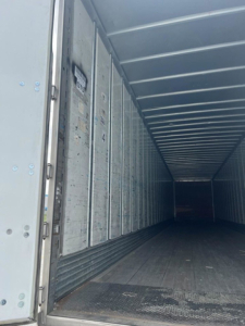 Interior of a trailer with metal sidewalls and a darkened long cargo area, entry from open rear door, visible riveted panels and floor.