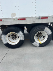 Trailer wheelset of a utility van showing dual rear axles with metal rims and tires; wheel hubs and lug nuts visible, part of a white refrigerated trailer side.