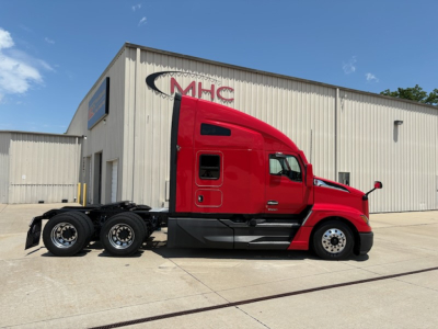 Kenworth T680 tractor in red color, side view, four rear wheels, parked in front of a beige industrial building with MHC sign.