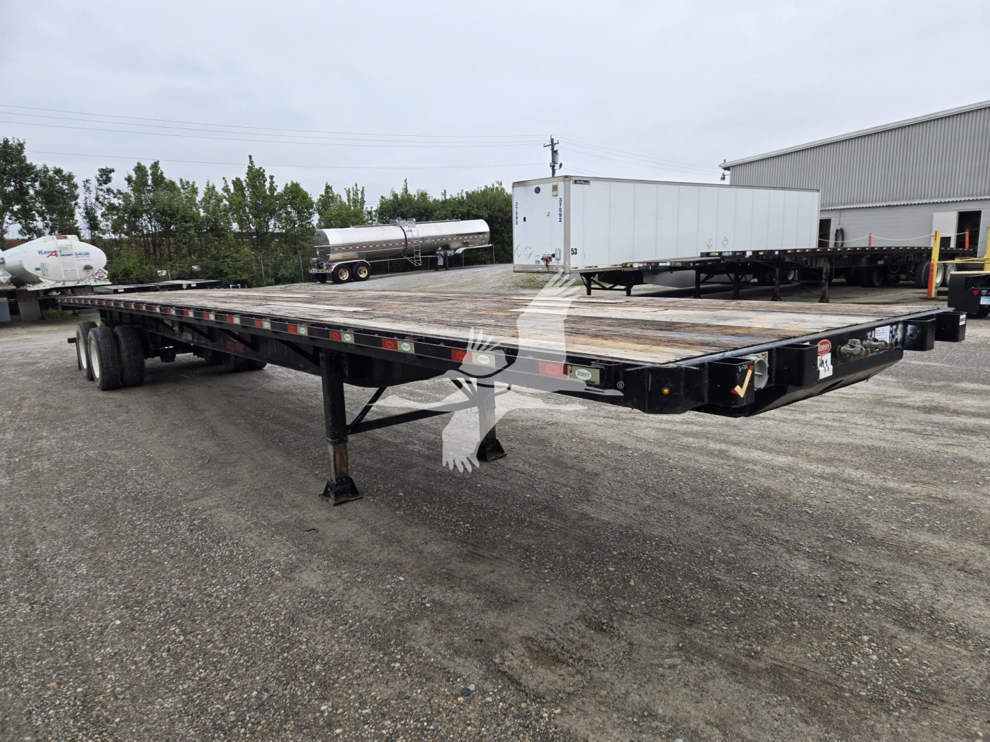 Flatbed trailer with dual rear axles, showing platform and supporting beams. Visible markings include small reflective tape along the edge and black frame. Industrial yard in background with other equipment.