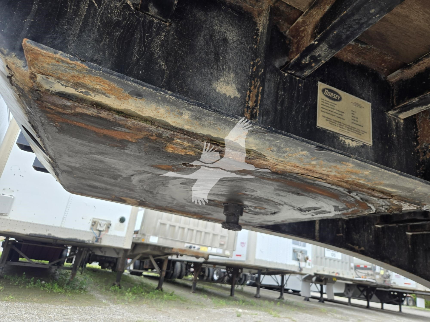 Flatbed trailer underside showing Dorsey plate and rust-streaked metal, with dirt and support beams.