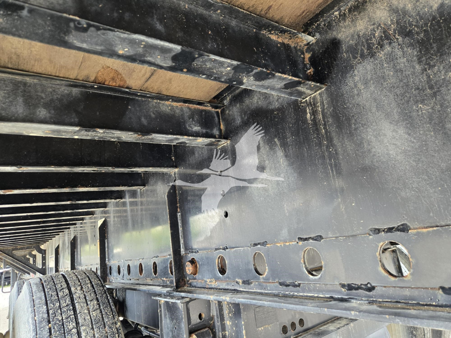Flatbed trailer undercarriage showing metal frame with riveted holes and wheel well area; black paint with dirt and reflections.