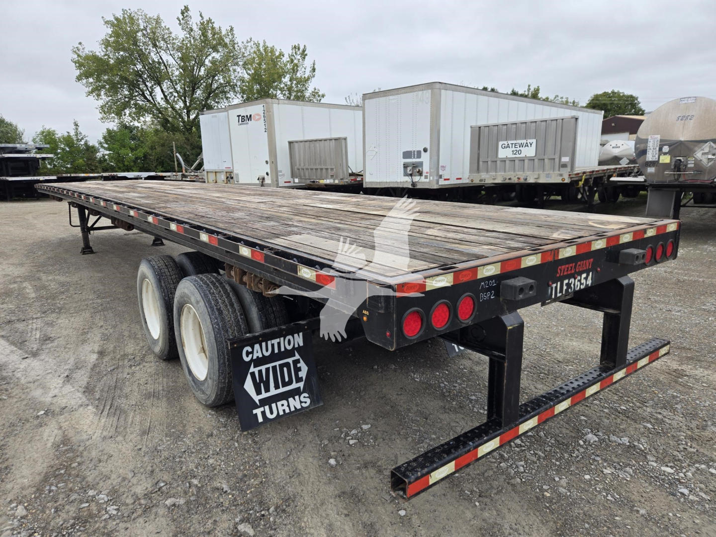 Trailer chassis with flatbed showing dual rear axles and taillights; CAUTION WIDE TURNS sign mounted on rear; worn wood deck; dock equipment and storage containers in background.