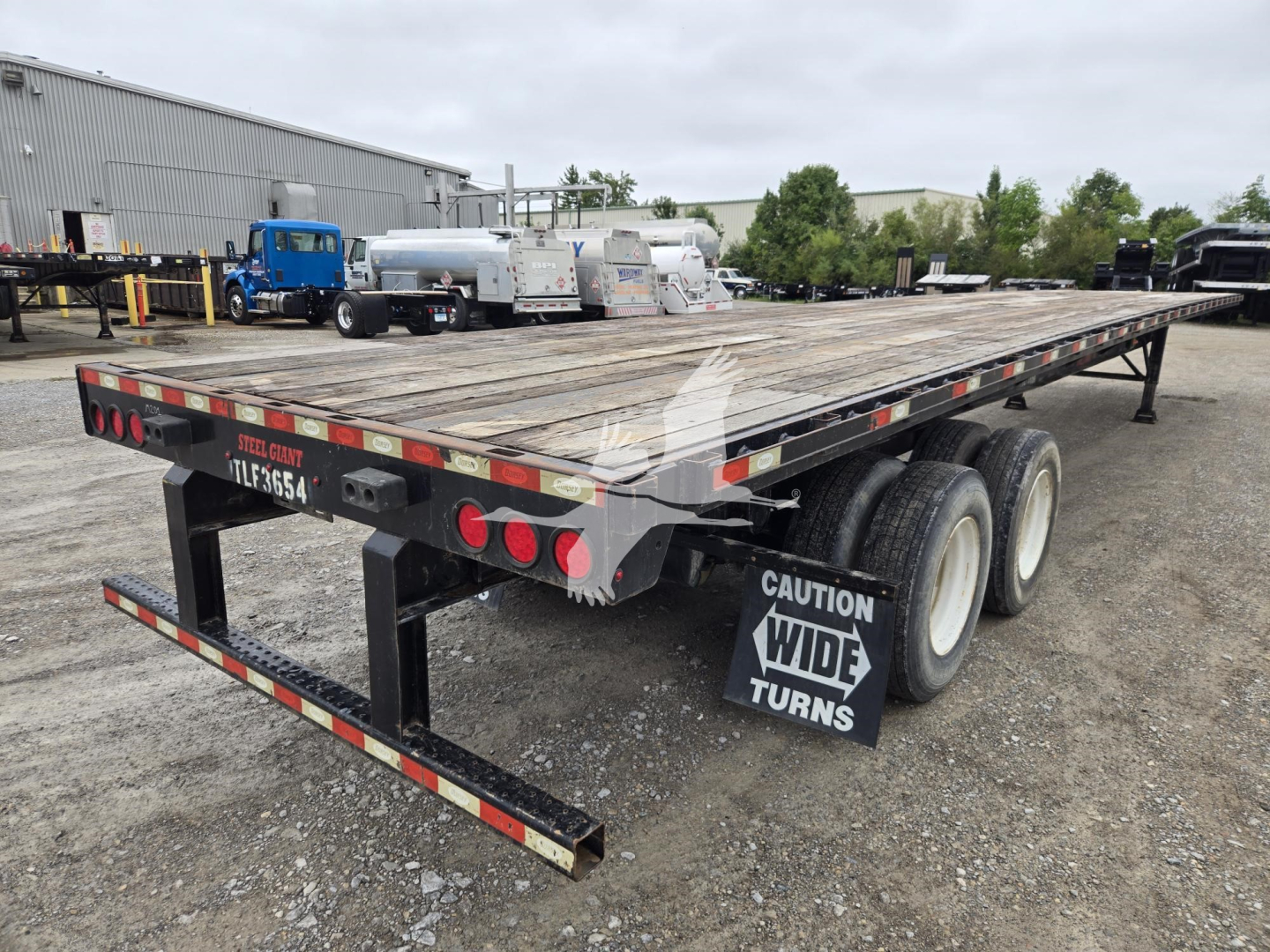 Flatbed trailer showing dual rear axles, wooden deck, and caution ‘Wide Turns’ mudflap; rear lights and license plate area visible.