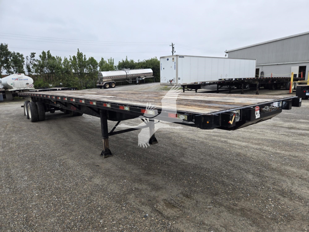 Flatbed trailer with dual rear axles, showing platform and supporting beams. Visible markings include small reflective tape along the edge and black frame. Industrial yard in background with other equipment.
