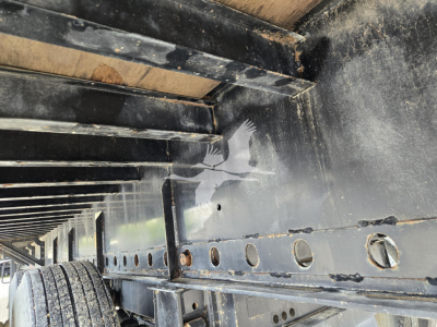 Flatbed trailer undercarriage showing metal frame with riveted holes and wheel well area; black paint with dirt and reflections.
