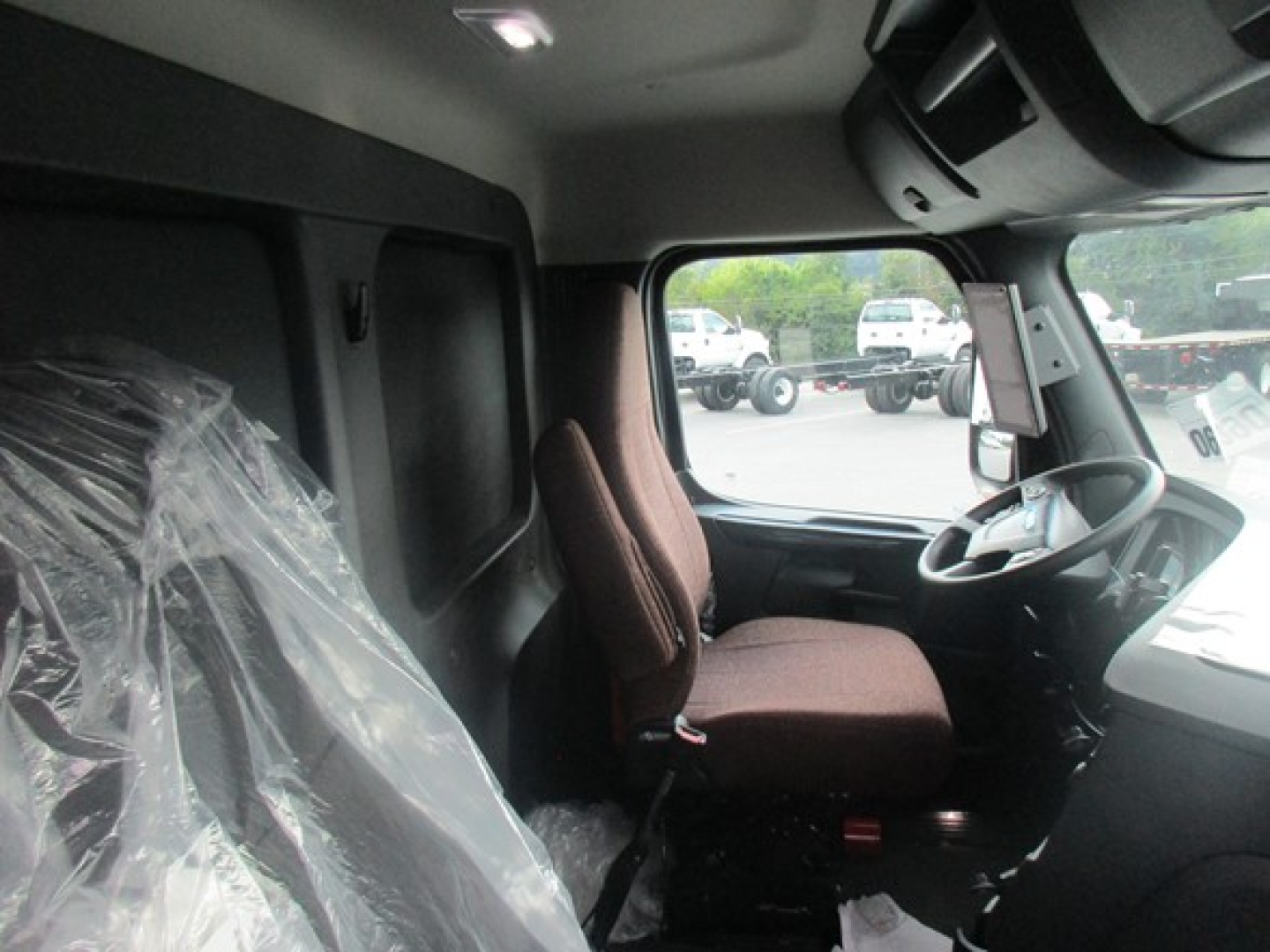 Truck cab interior showing steering wheel and driver seat in a Freightliner Cascadia 126, with plastic-covered seat on left and dashboard visible.