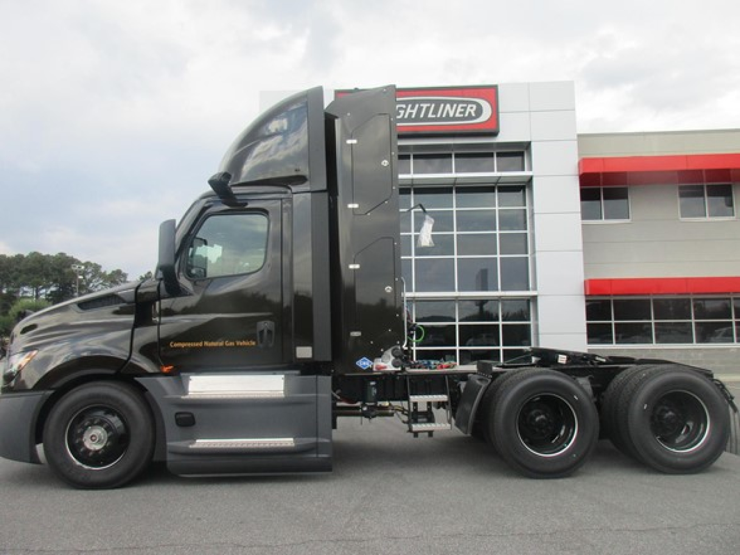 Semi truck cab and chassis in front of Freightliner dealership building. Door area shows text; may include branding and model details.