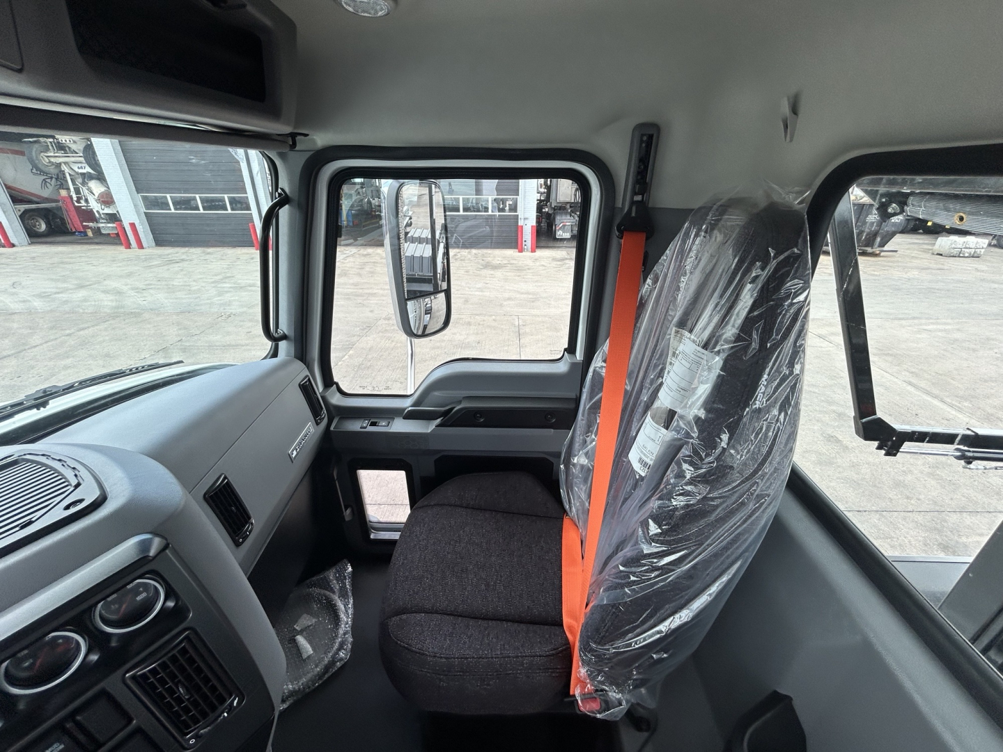 Cab interior of a 2027 Mack GR64B roll-off truck showing driver seat with plastic wrapping, orange seat belt, dashboard, and side window.