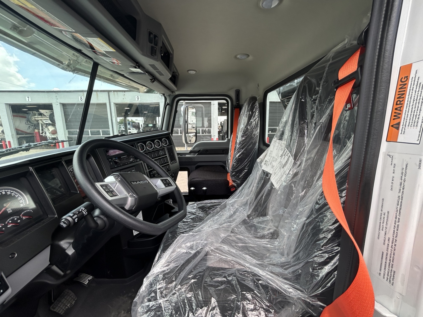 Cab interior of a 2027 Mack GR64B roll-off truck showing steering wheel, dashboard, and wrapped seats with plastic covers.