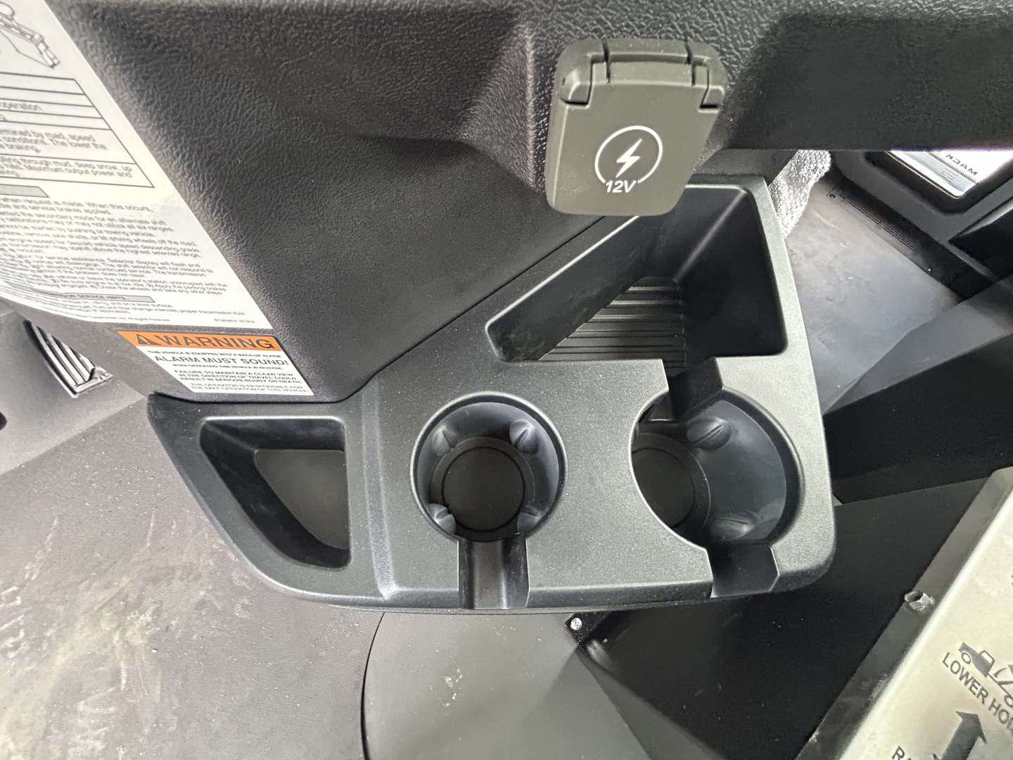 Dashboard area of a Mack GR64B showing two cup holders and a 12V power outlet cover with 12V icon.
