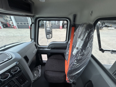Cab interior of a 2027 Mack GR64B roll-off truck showing driver seat with plastic wrapping, orange seat belt, dashboard, and side window.