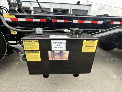 Truck chassis with a hydraulic oil reservoir tank mounted, showing labels: CAUTION, PRECAUCION, HYDRAULIC OIL ONLY CAP 40 GAL, and sticker with GH logo; black metal box.