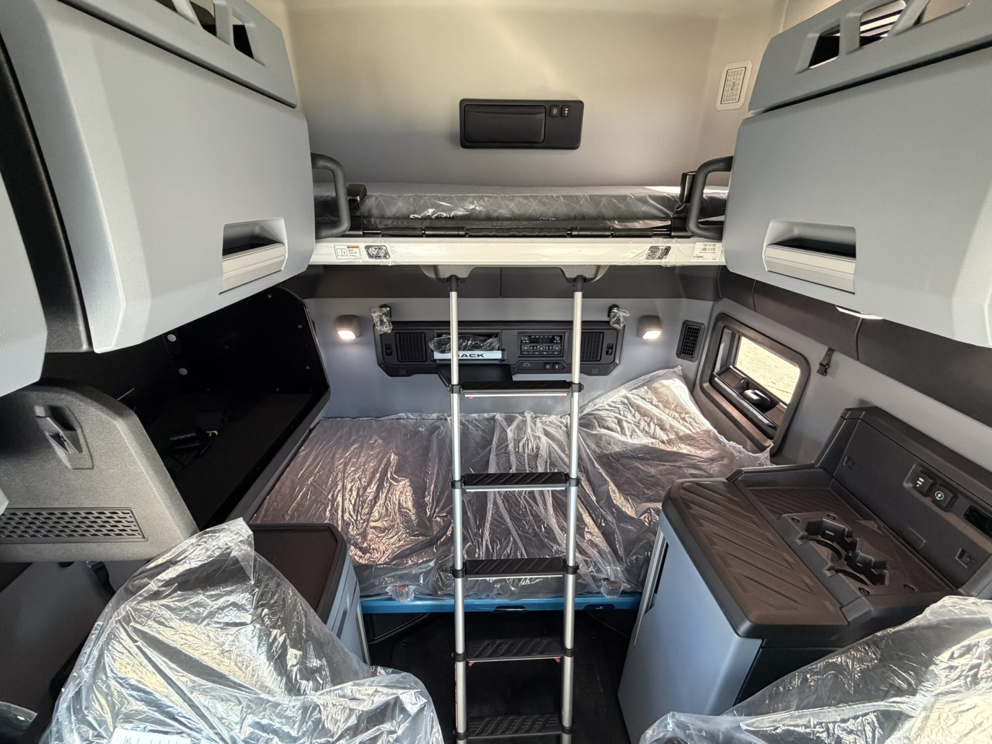 Mack PR64T76H cab interior showing bunk bed area with ladder, overhead storage cabinets, and command center in front; plastic-covered flooring and seating.