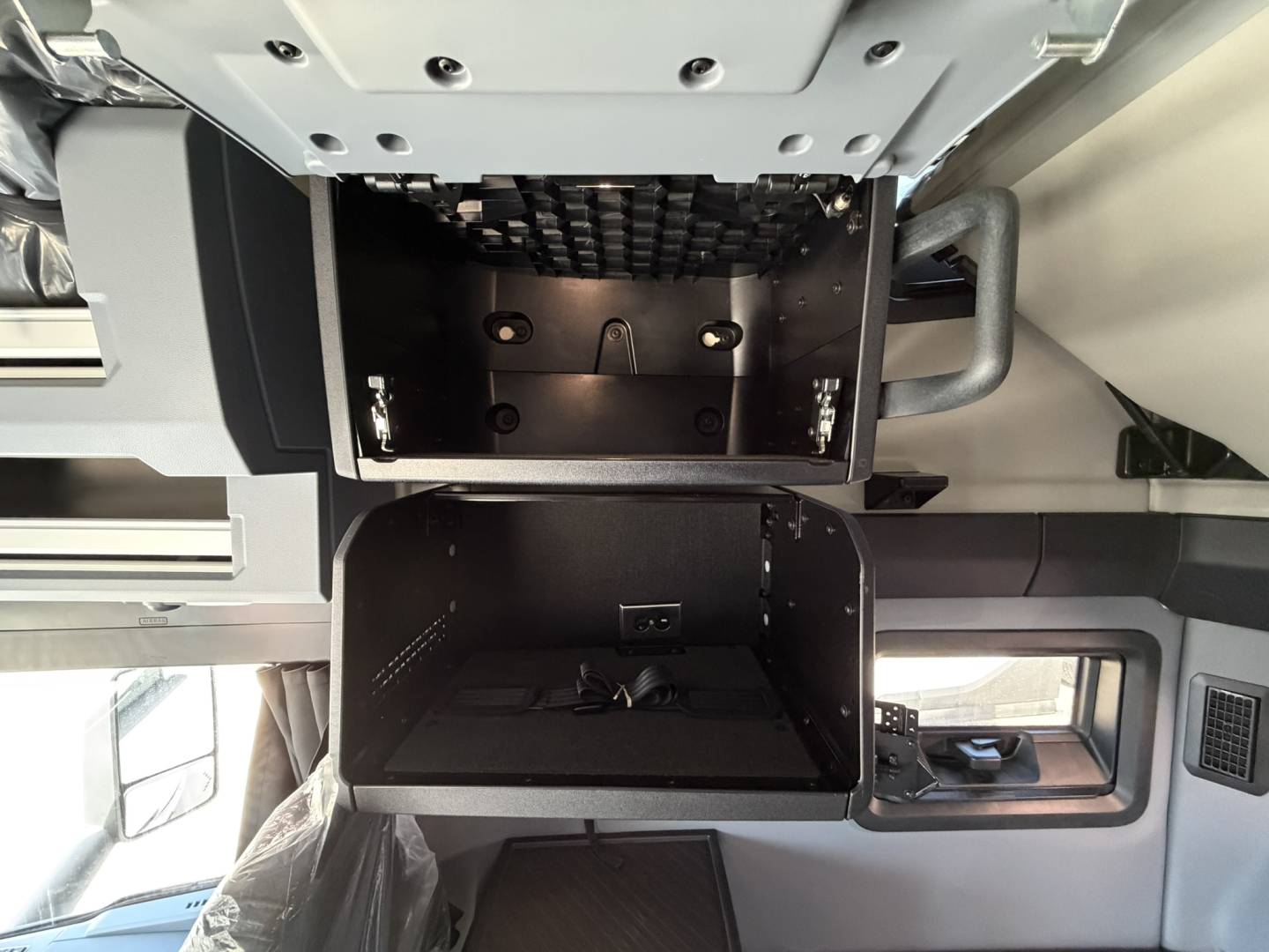 Truck cab interior showing two open bays/lockers under the dash area in a Mack PR64T76H with plain panels and mounting hardware visible.