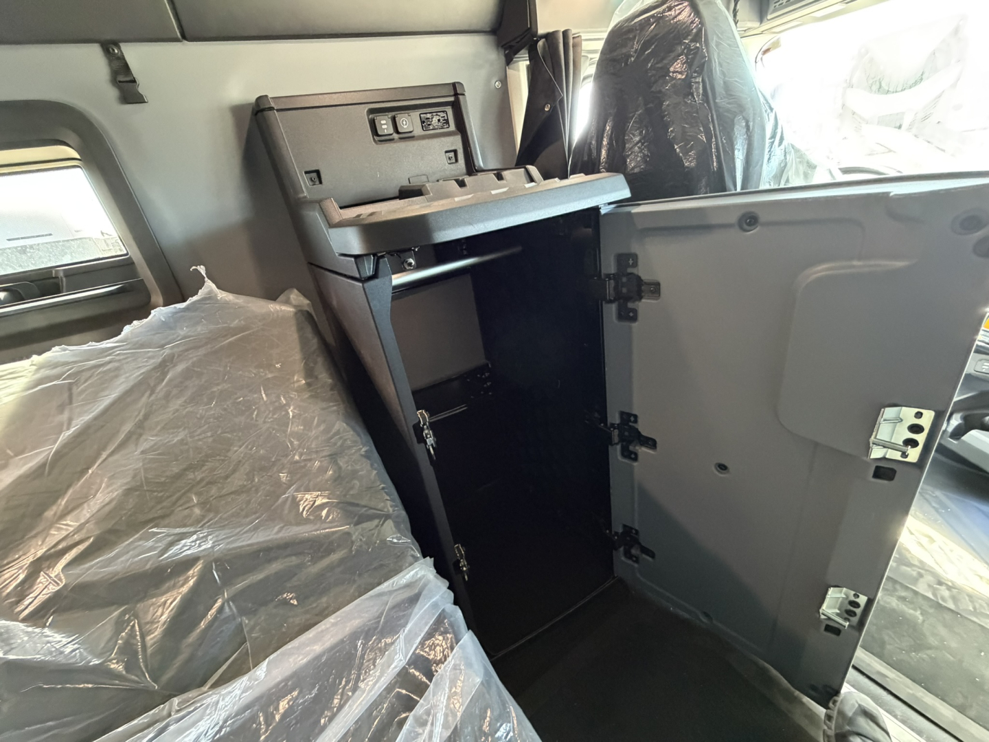 Truck cabin interior showing open cabinet/storage compartment and seating area with plastic-covered seats; badges and controls visible near door.