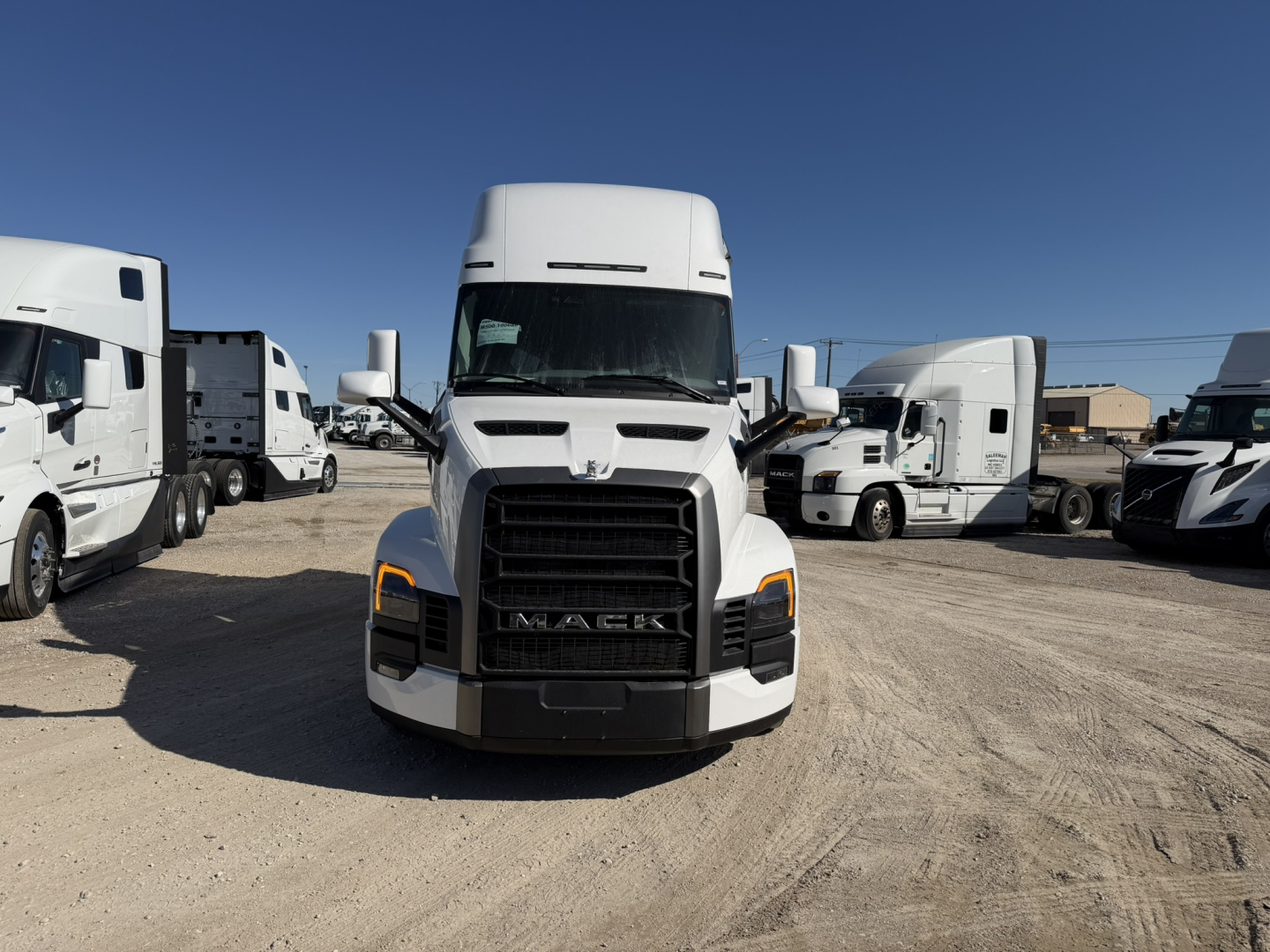 Front view of a white 2026 Mack PR64T76H truck in a lot, with other semi-trucks in background, showing grille and headlights.