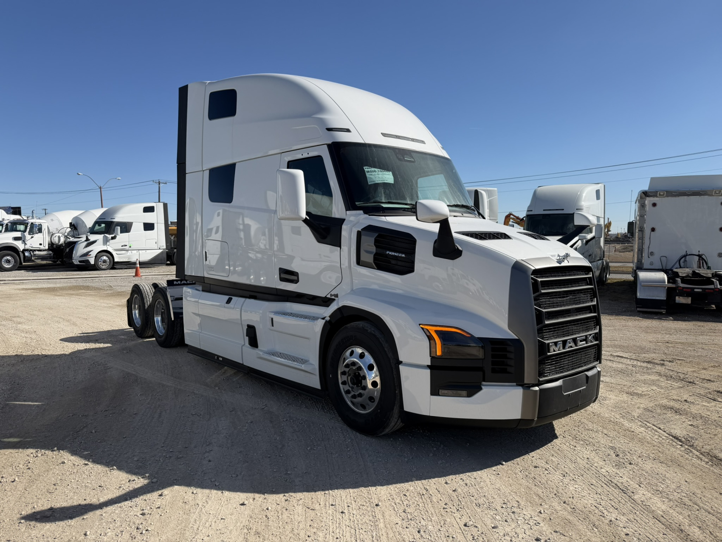 Truck cab in a dealership lot showing a 2026 Mack PR64T76H sleeper with cog wheel grille, white exterior, chrome bumper, and dual rear axles.