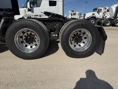 Two rear axles of a Mack PR64T76H truck chassis with dual tires, chrome rims, and black undercarriage; gravel lot and other trucks in background.