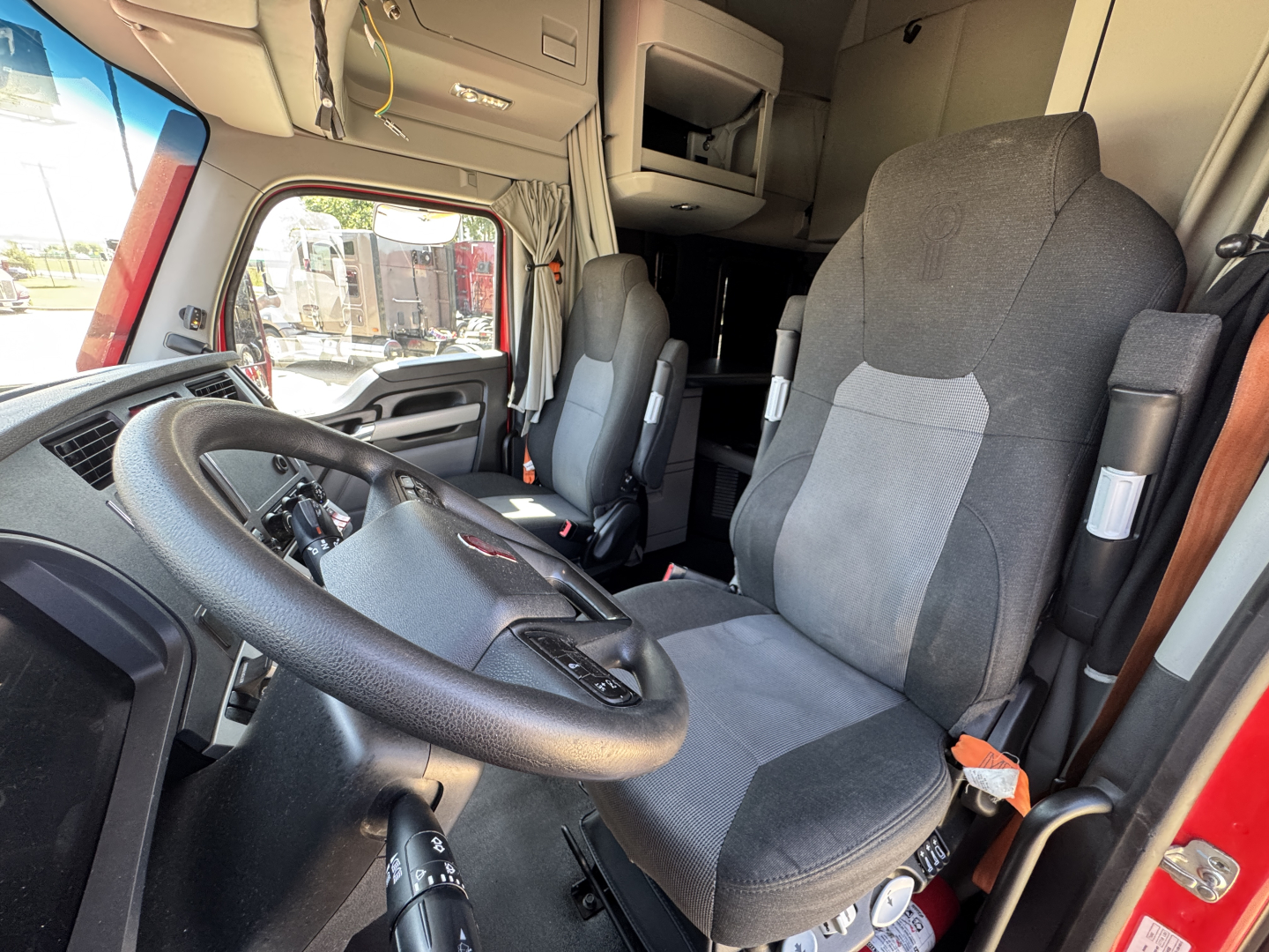 Cab interior of Kenworth truck showing steering wheel, driver seat, dashboard, and bunk area with overhead storage.