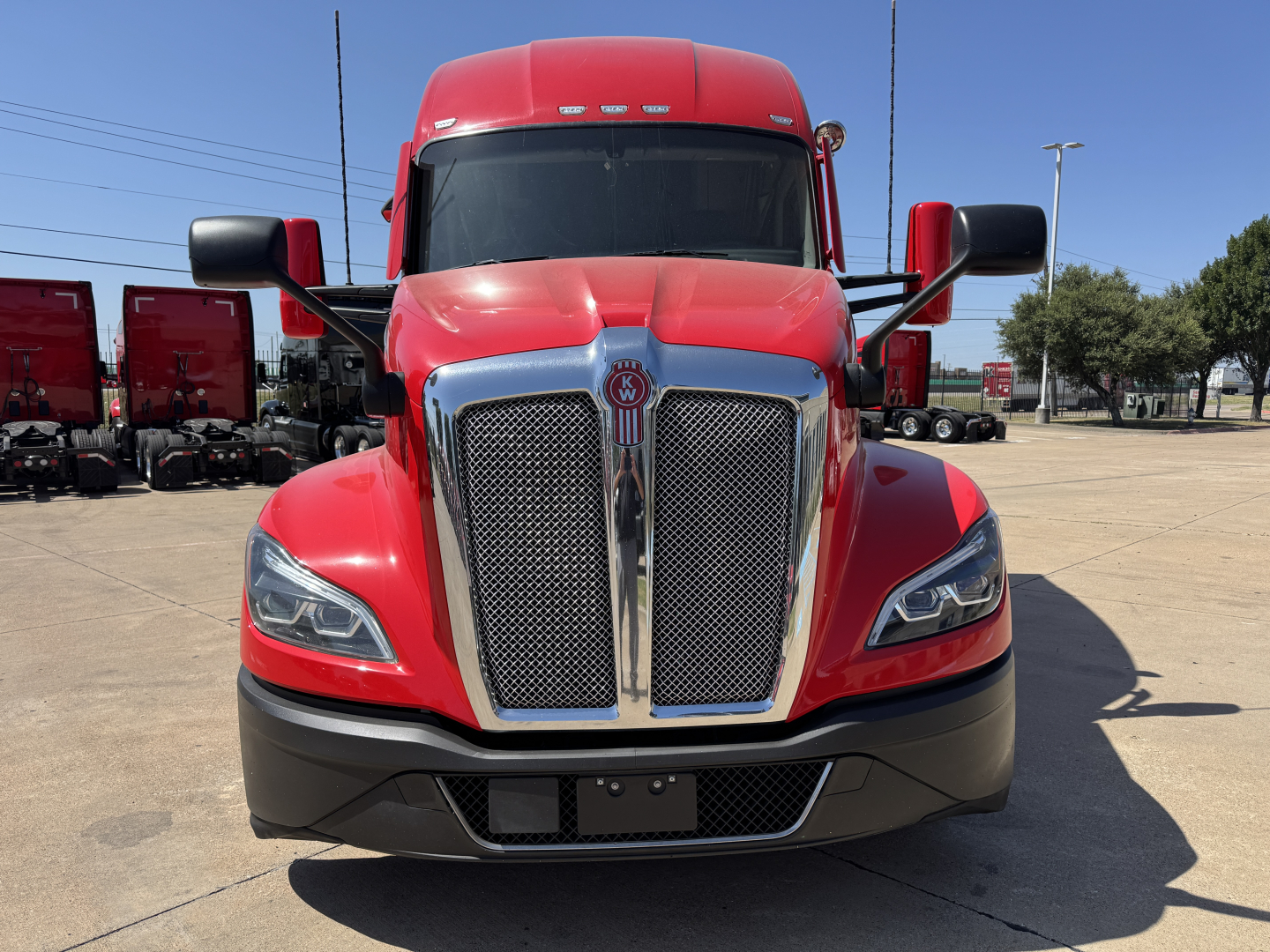 Kenworth T680 front view with red cab, chrome grille, black lower bumper, daytime highway setting.