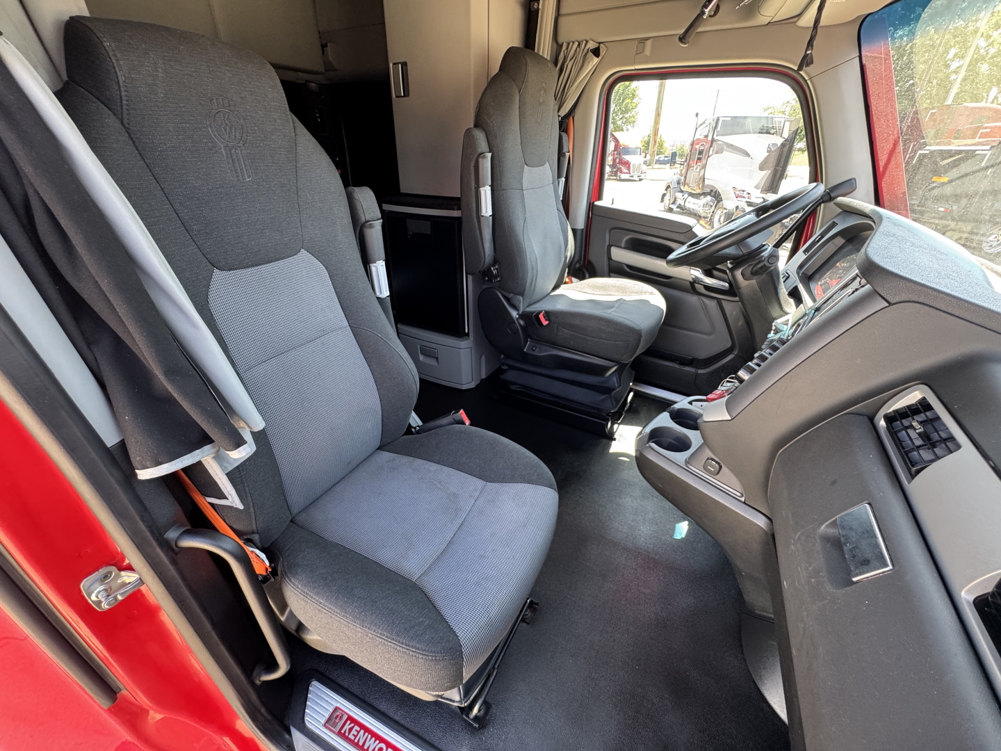 Kenworth T680 truck interior driver and passenger seats visible with grey fabric, dashboard, steering wheel, and open cab entry.