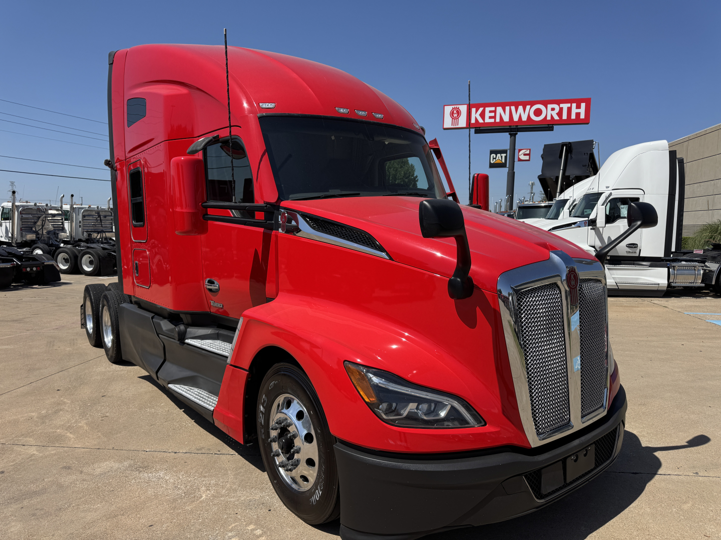 Kenworth T680 sleeper cab truck in red, front three-quarter view, parked at dealership lot with Kenworth sign in background.