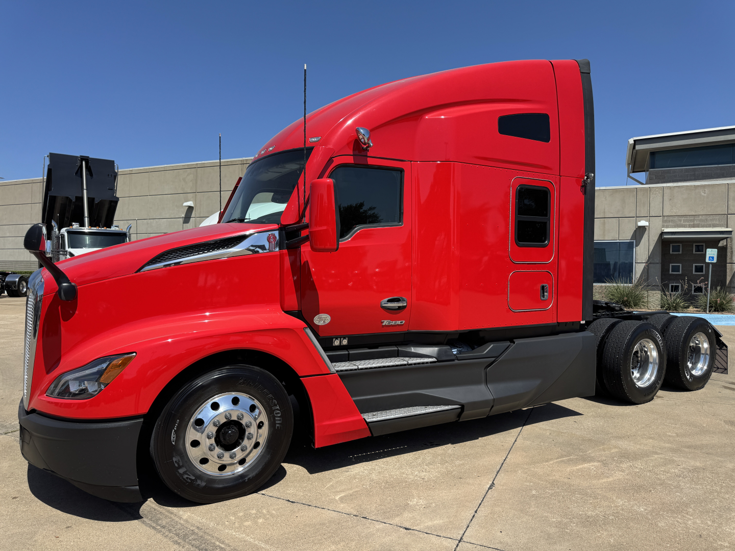 Red Kenworth T680 conventional sleeper tractor, side view with fuel tanks and chrome wheels; no discernible labels or stickers visible on the cab from this angle.