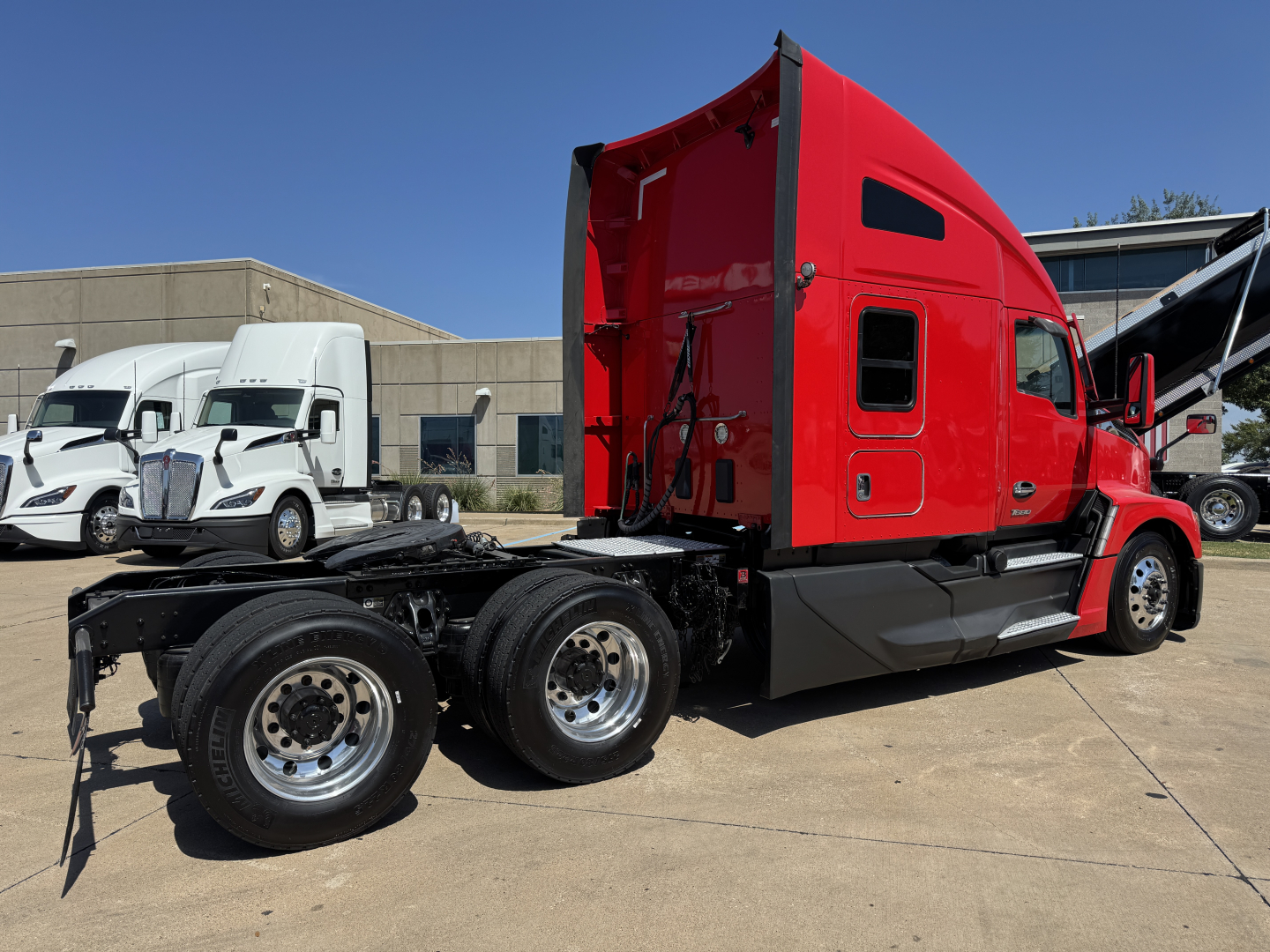 Kenworth T680 truck chassis side view showing cab with red paint and sleeper; rear wheels and fifth wheel visible; other white trucks in background.