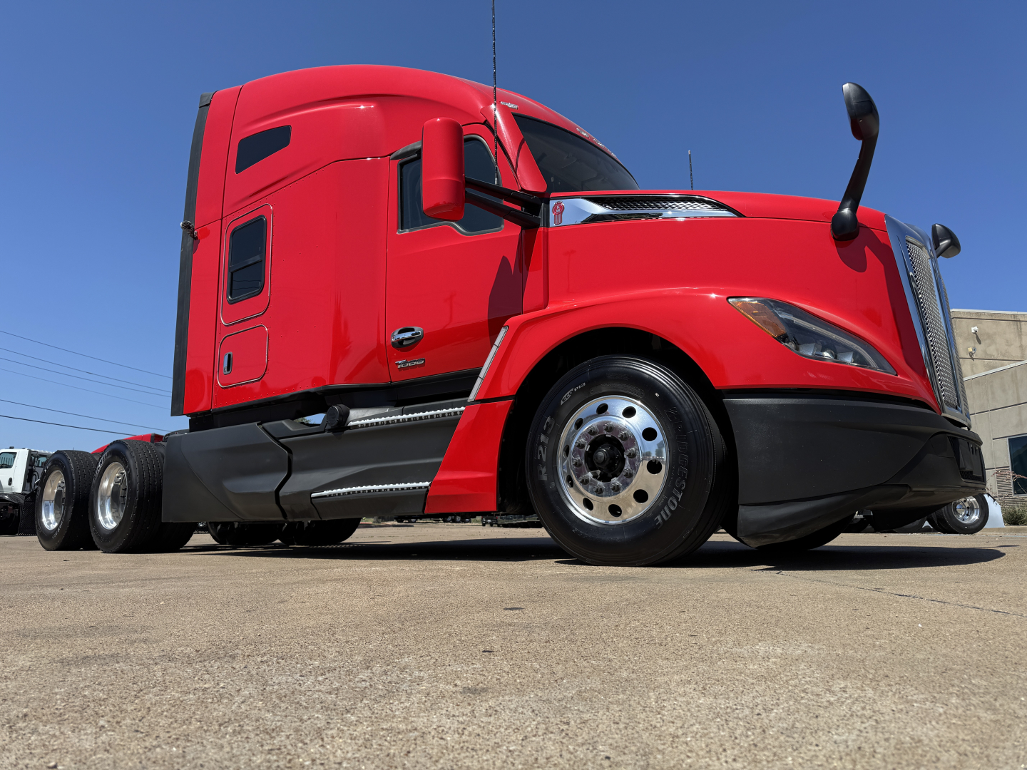 Kenworth T680 tractor-trailer side view in red showing front wheel, fuel tanks, and sleeper cab with windows; no legible text visible on the truck in this image.