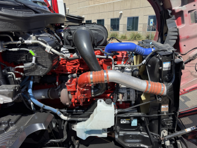 Kenworth T680 engine bay showing red engine block, blue silicone coolant hose, black air intake, silver exhaust pipe, and radiator with coolant reservoir visible.