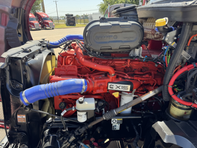 Kenworth T680 engine bay showing red Cummins ISX engine with blue hoses and black plastic covers; various components including turbo, hoses, and coolant reservoir visible.