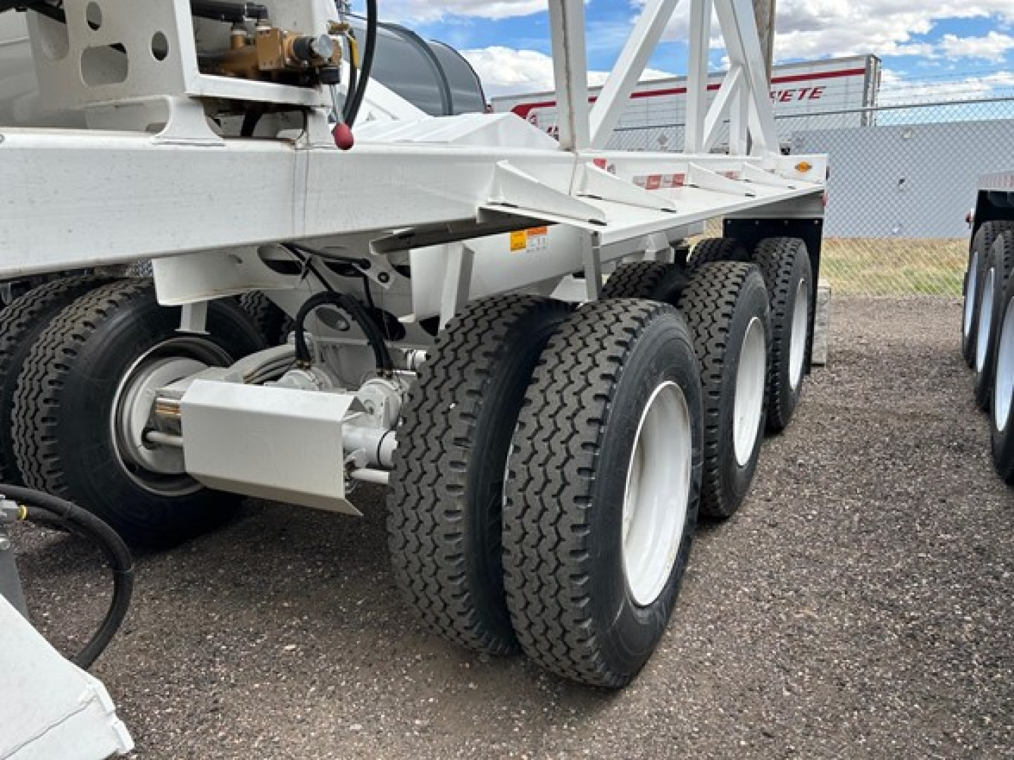 Trailer chassis with tandem axles and large dual tires, white frame, visible hydraulic hoses and equipment mountings on left side.