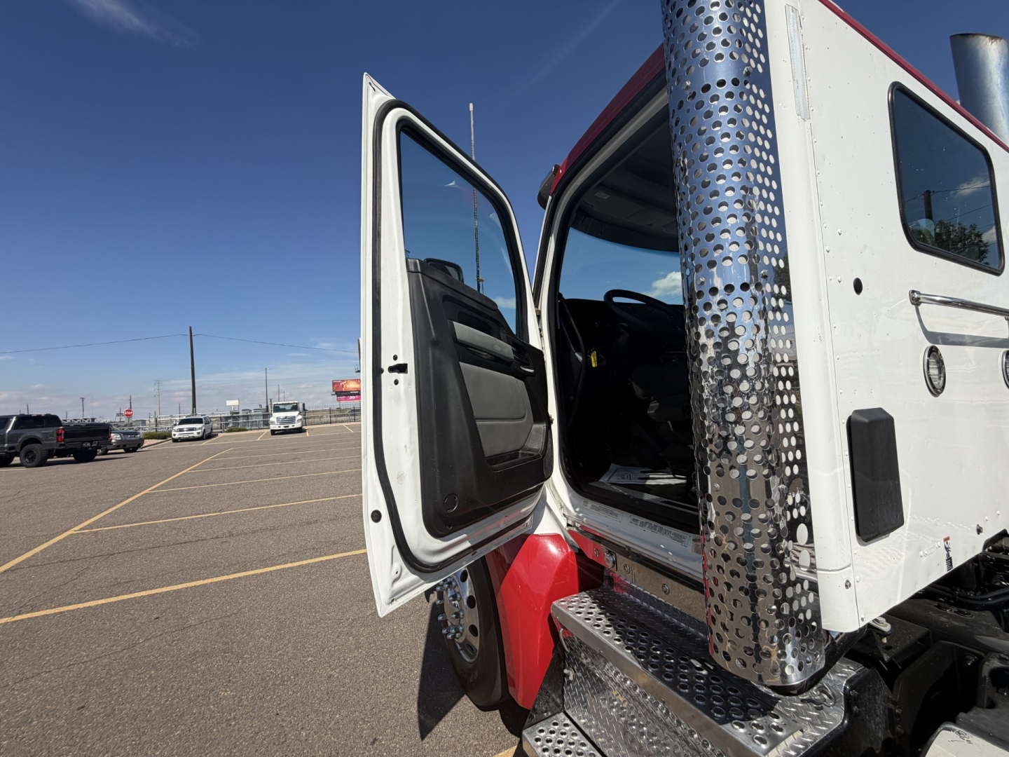 Kenworth T880 cab exterior with open driver door showing step, chrome perforated metal side panel, and white cab; truck in lot.