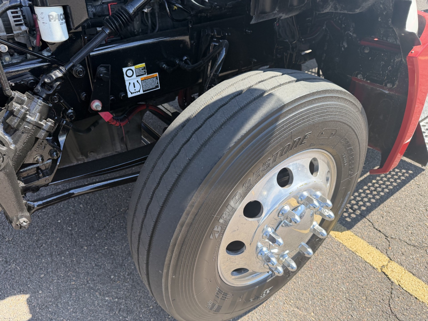 Kenworth T880 front axle and wheel assembly showing large dual-tire on front? but single steer axle wheel with polished hub and lug nuts; surrounding chassis and hydraulic components visible.