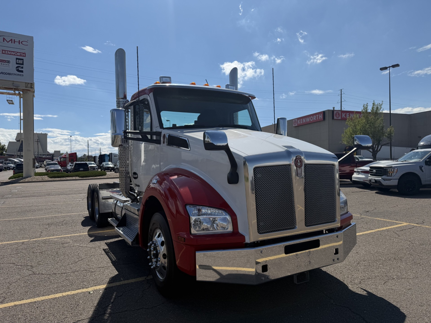 Kenworth T880 conventional daycab truck in a dealership lot, front angled view, large chrome grille, dual exhaust stacks, red fenders, chrome bumper, white cab.
