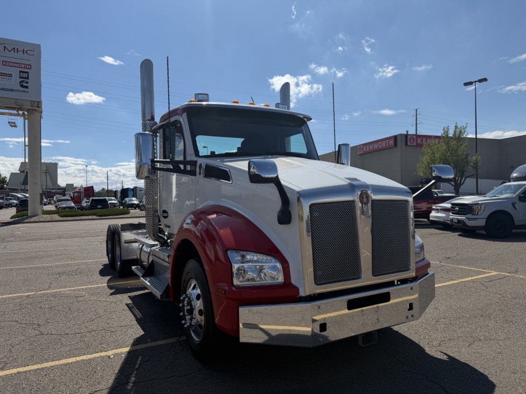 Kenworth T880 conventional daycab truck in a dealership lot, front angled view, large chrome grille, dual exhaust stacks, red fenders, chrome bumper, white cab.