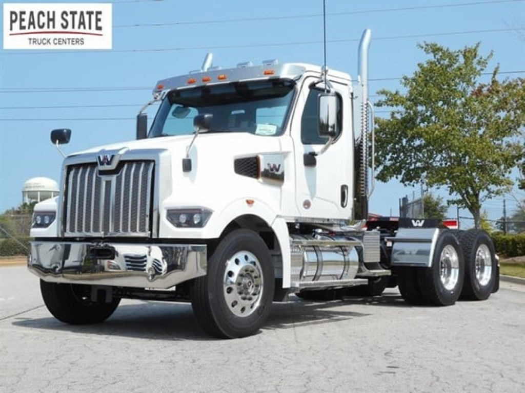 Front-left view of a white 2026 Western Star 49x day cab truck showing chrome grille, dual exhaust stacks, and manufacturer graphics.