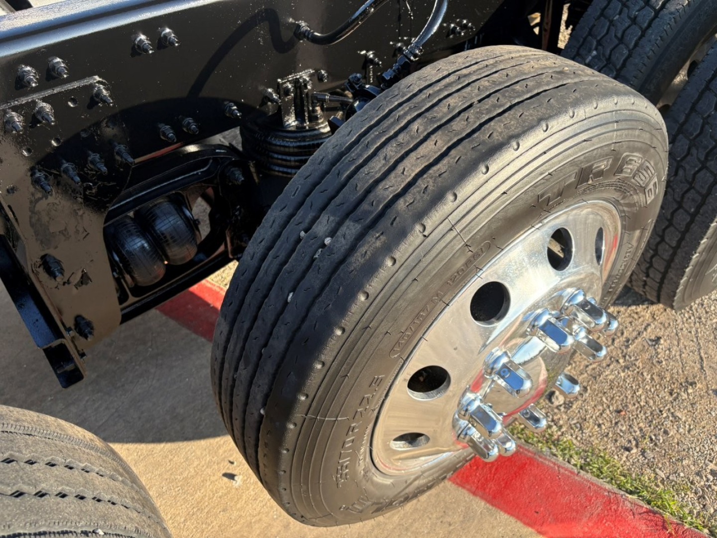 Truck front wheel assembly showing dual rear axle leaf springs and tire with Volvo logo hub, chassis, and hydraulic components in view.