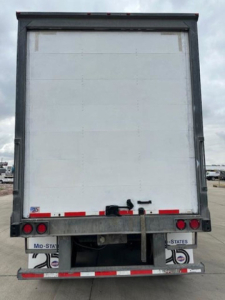 Rear view of a white WB-type trailer with a metal frame and red reflectors. No visible text legible on the door; taillights and bumper seen.