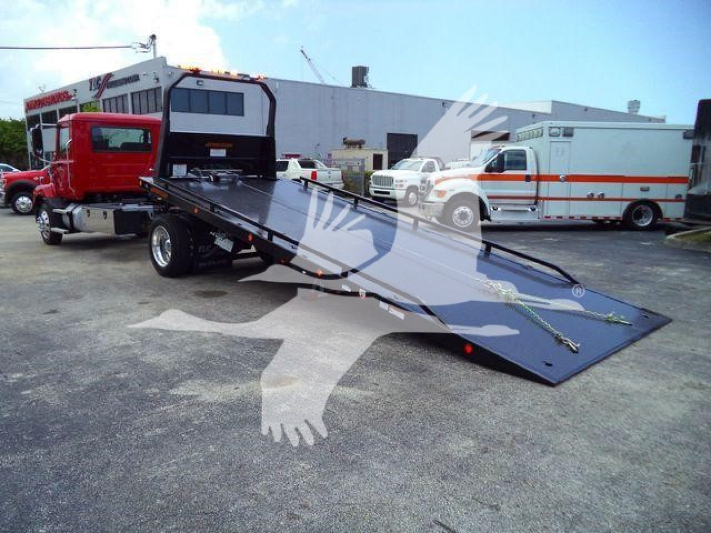MD6 tow truck with flatbed in raised position, red truck in foreground, emergency vehicles in background.