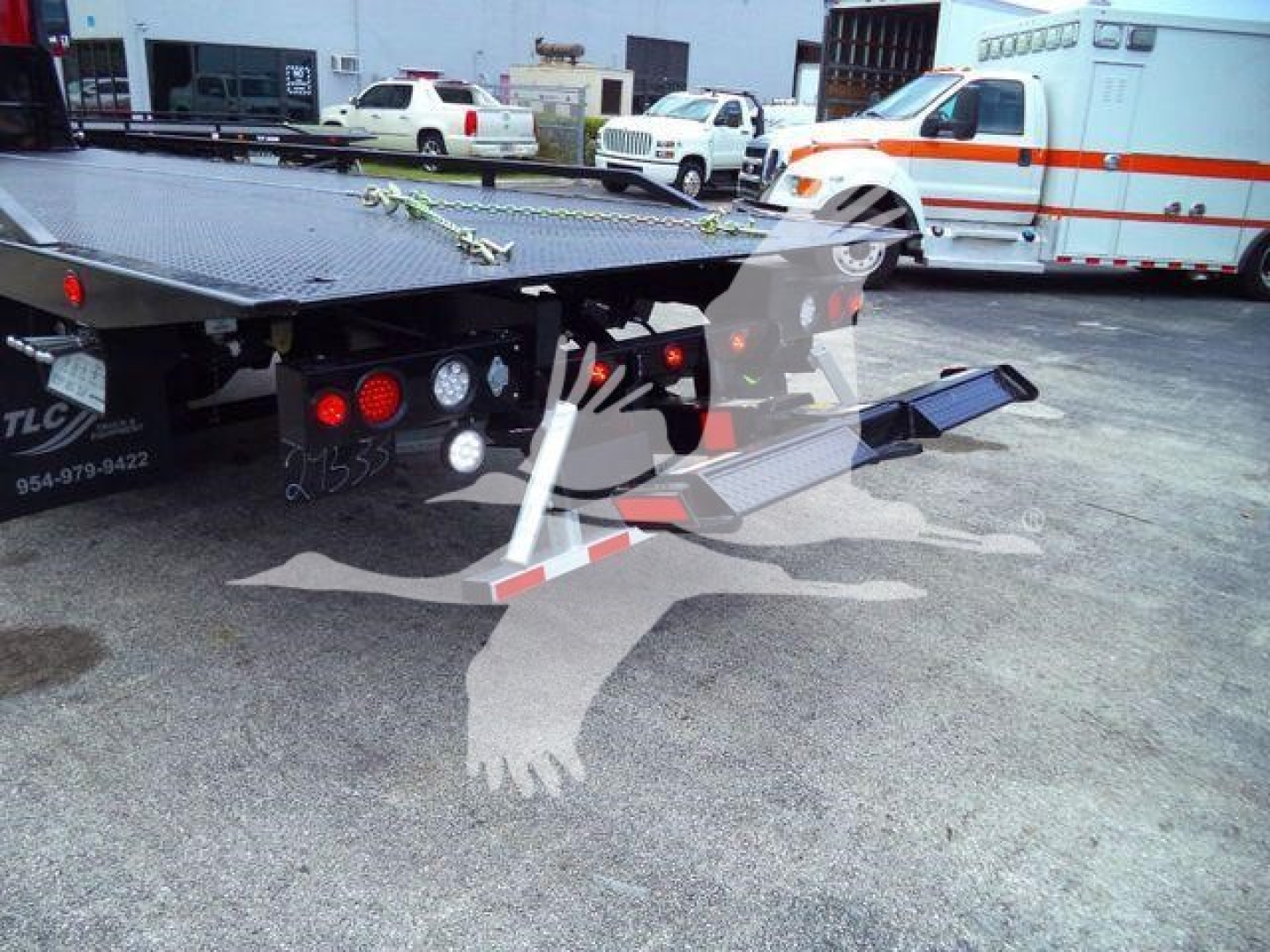 Flatbed tow truck with ramp extended showing rear view, emergency vehicle in background, and various trucks in lot.