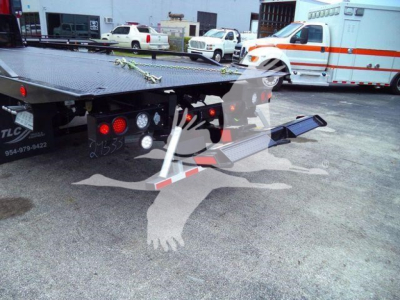 Flatbed tow truck with ramp extended showing rear view, emergency vehicle in background, and various trucks in lot.
