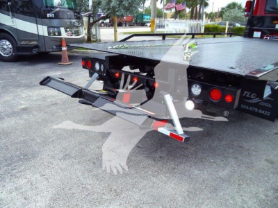 Tractor-trailer rear view showing a rollback bed with two fold-down ramps and taillights; hydraulic system and storage area visible under bed.